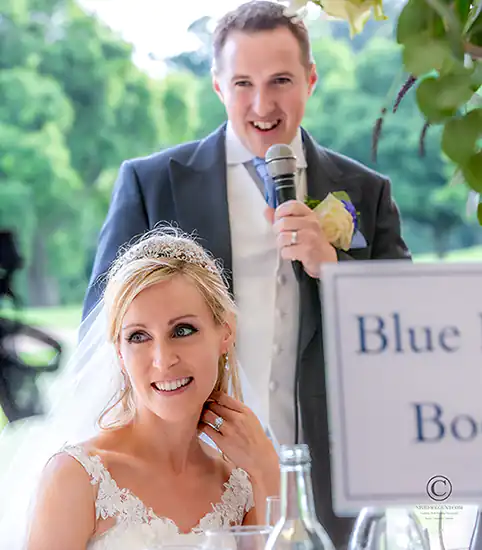 Groom delivers wedding speech in grounds of Ashdown Park Hotel near Tunbridge Wells as bride listens with a smile