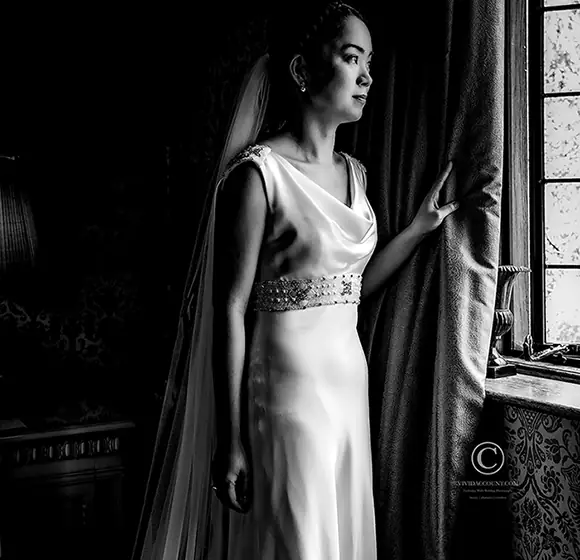 Bride in slim, silk and jewelled wedding dress takes a moment to gaze out of the window of the Bridal Suite at Langshot Manor before her wedding starts