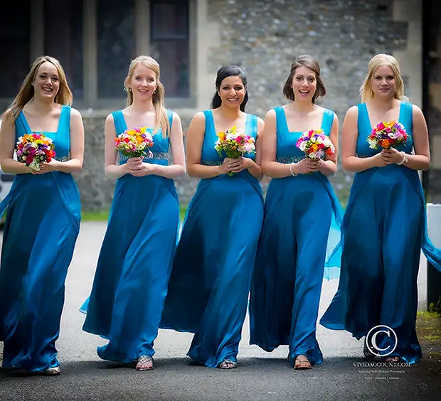 5 bridesmaids in full length blue dresses holding colourful tunbridge wells bouquets walk side by side towards church for start of wedding