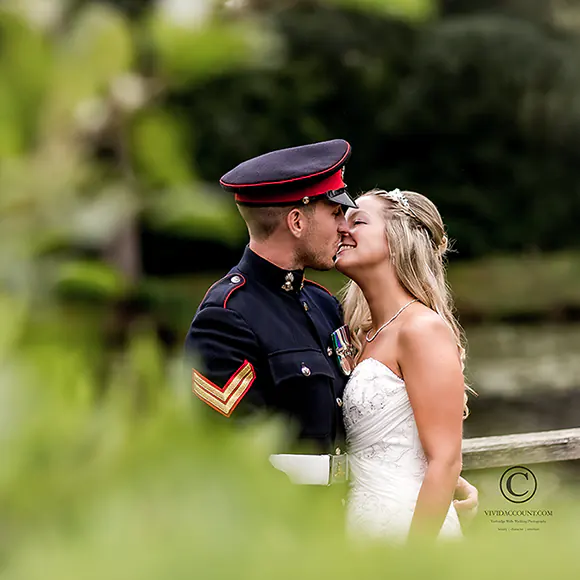 Military groom gives his new wife a wedding day kiss partially hidden behind foliage in the grounds of Ashdown Park Hotel, near Tunbridge Wells, Kent